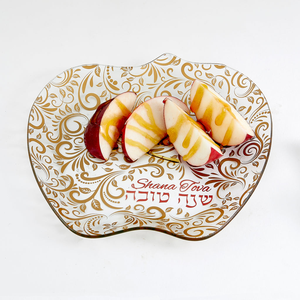 An apple-shaped glass serving platter with a white and gold floral pattern, featuring the words 'Shana Tova' and Hebrew text, shown with sliced apples on a white background.