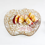 An apple-shaped glass serving platter with a white and gold floral pattern, featuring the words 'Shana Tova' and Hebrew text, shown with sliced apples on a white background.