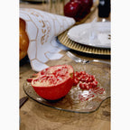 A halved pomegranate with exposed seeds and a small pile of loose pomegranate seeds on a clear glass plate with gold ornamental detailing, set on a wooden table with plates and a napkin in the background.