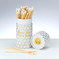 A white cylindrical match holder with a gold honeycomb pattern, filled with wooden matches with white tips. The front of the holder reads 'Shana Tova' in English and Hebrew, and the lid lies beside it showing a gold menorah logo and 'The Jacob Rodriguez Matcha Collection' text.