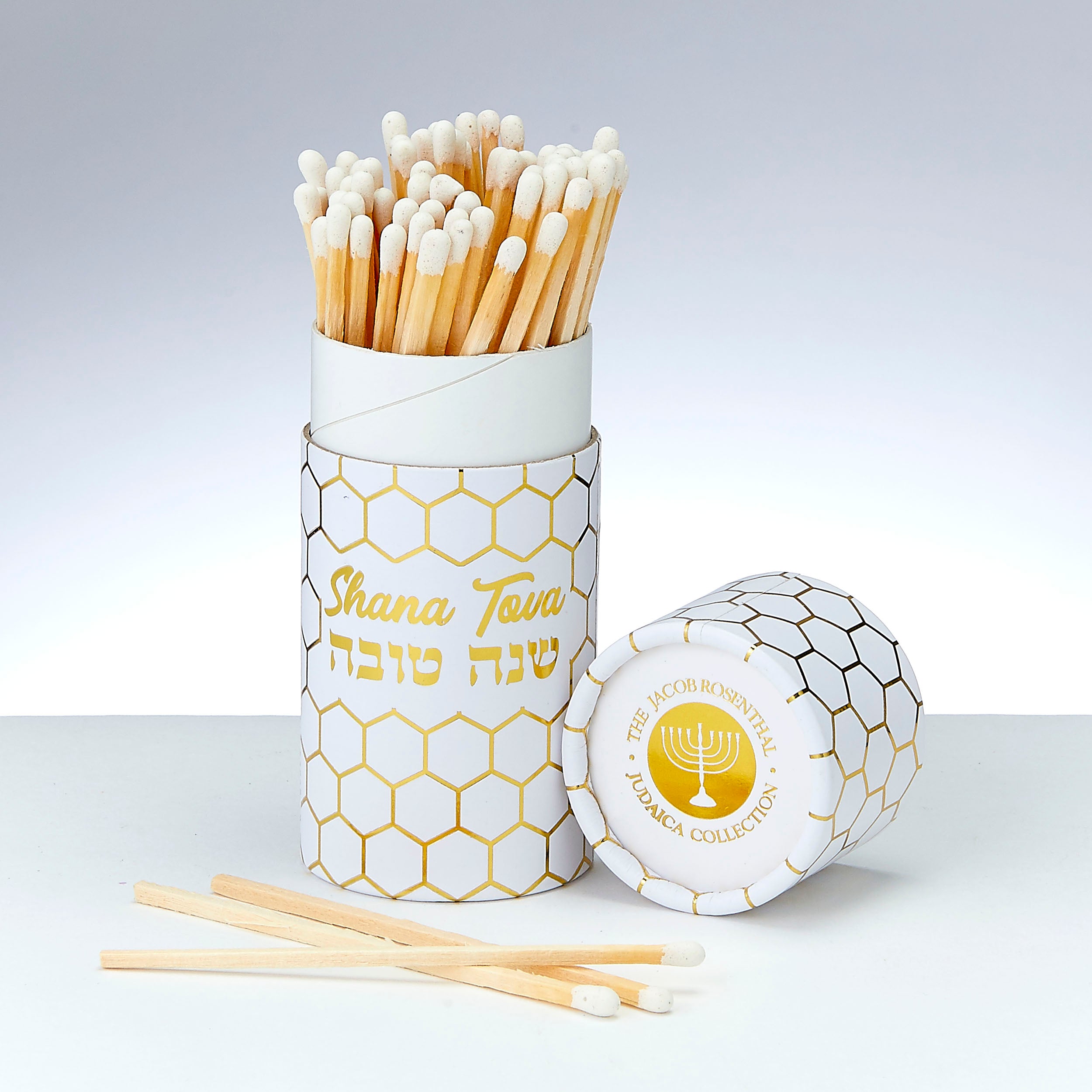 A white cylindrical match holder with a gold honeycomb pattern, filled with wooden matches with white tips. The front of the holder reads 'Shana Tova' in English and Hebrew, and the lid lies beside it showing a gold menorah logo and 'The Jacob Rodriguez Matcha Collection' text.