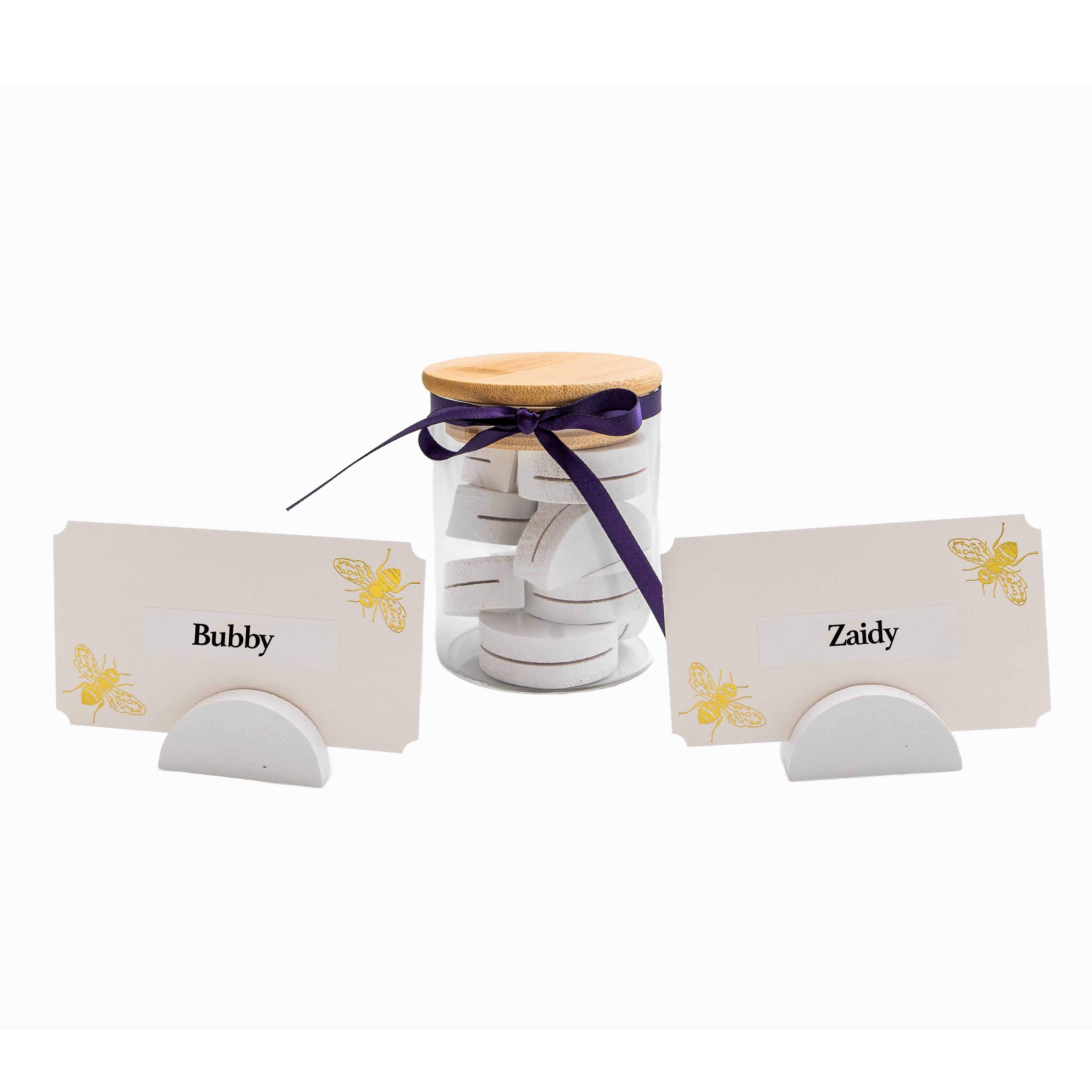 A set of personalized wooden name cards for guests, shown with two cream-colored cards labeled 'Bubby' and 'Zaidy' with gold bee illustrations, and a clear glass jar filled with round white wooden tokens tied with a dark purple ribbon and topped with a light wood lid.