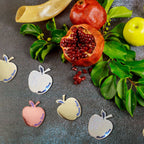A styled flat-lay of metallic apple-shaped nameplates with the text 'Moshi Tza'at' in blue script, arranged on a dark textured surface with pomegranate halves, green leaves, a yellow apple, and a brass shofar.