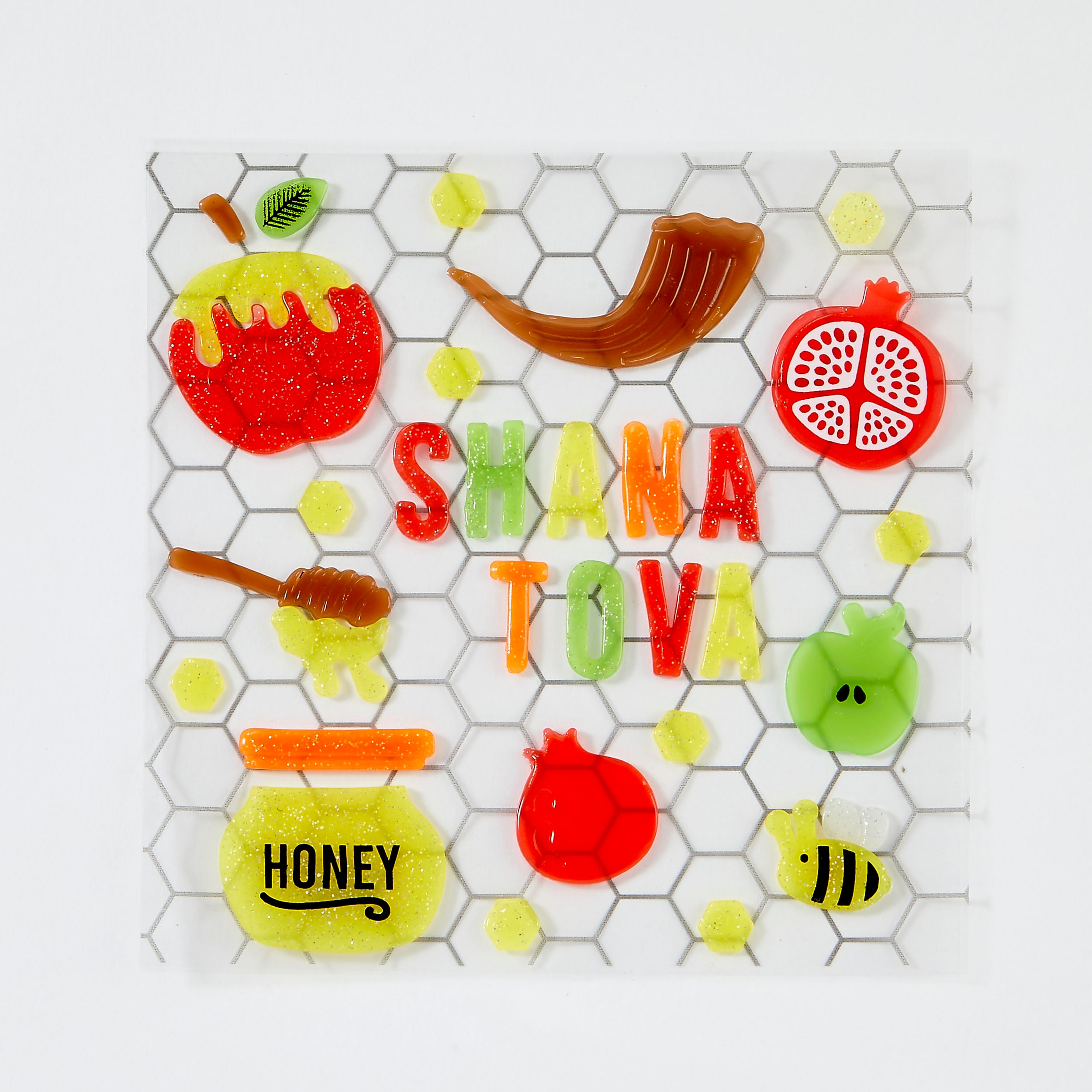 A square with stickers featuring a honeycomb pattern and the Hebrew text 'Shana Tova' in colorful letters, surrounded by festive honey-themed illustrations including a jar of honey, a pomegranate, a honey dipper, a bee, and a dripping red apple.