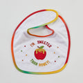 A white baby bib with a rainbow-colored stitched border and an embroidered design featuring a red apple with honey dripping from it, surrounded by the text 'I'M SWEETER THAN HONEY!'.