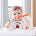 A baby sitting in a white high chair, holding an orange spoon near their mouth while wearing a white bib with a red apple graphic and the text 'I'M SWEETER THING.