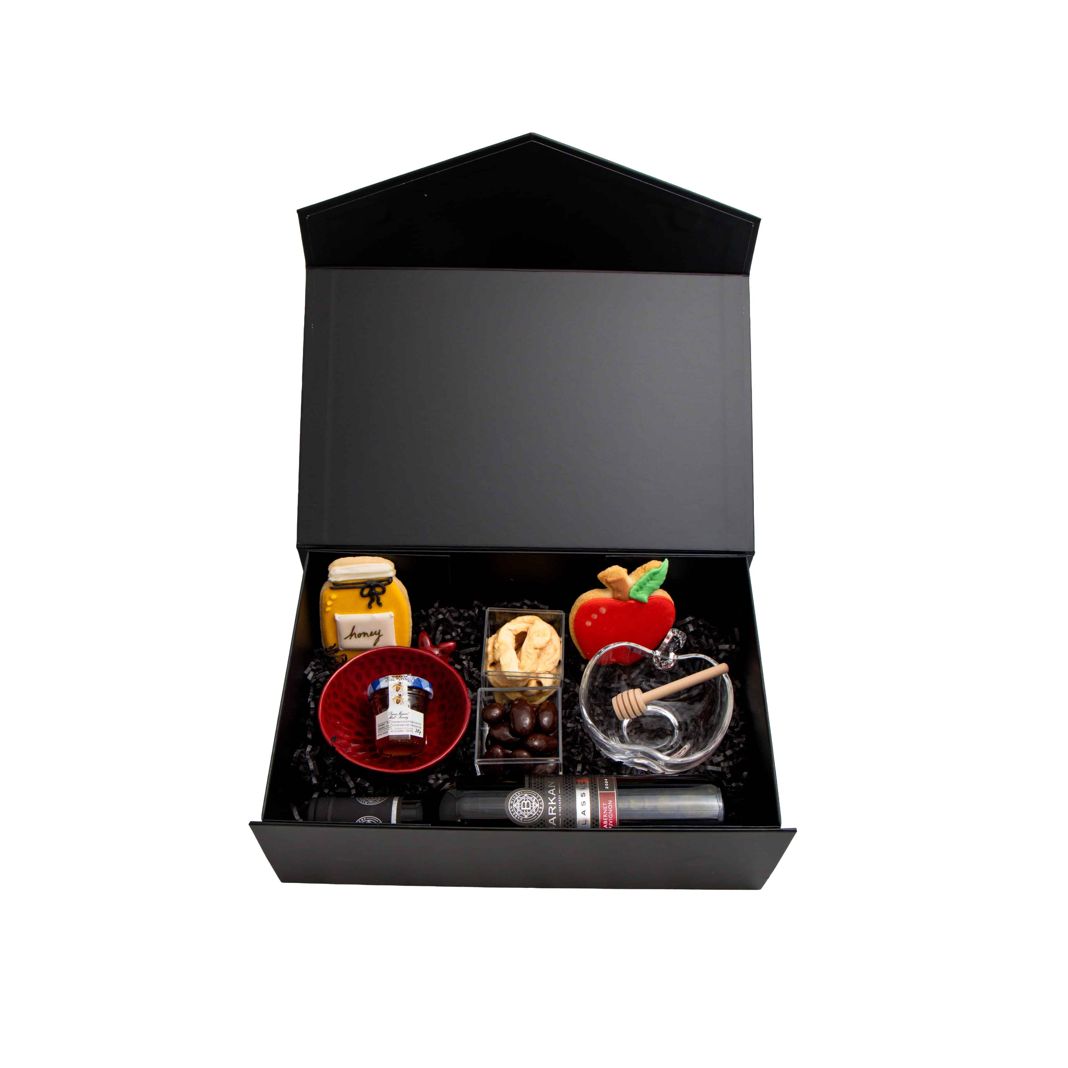 A black gift box with a house-shaped lid, shown open on a white background. Inside are several small gift items, including a small bottle, a red bowl, a glass with a cinnamon stick, a small jar with a red apple lid, and other confectionery or accessory items arranged in black filler.