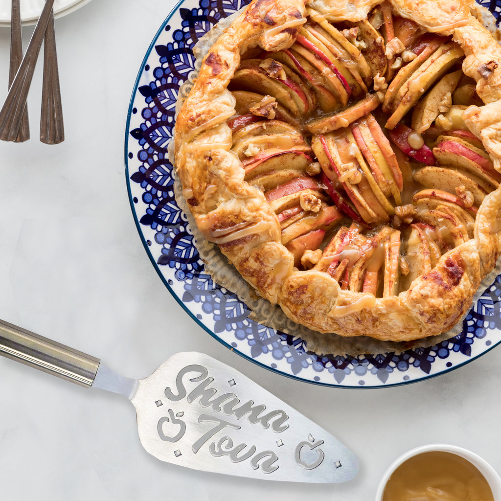 A baked apple pie with a flaky crust and sliced apples sits on a blue-and-white patterned plate on a light marble surface. A silver serving spatula engraved with the name 'Shana Tova' is placed beside the plate, along with a small cup of coffee and the corner of a wooden spoon.