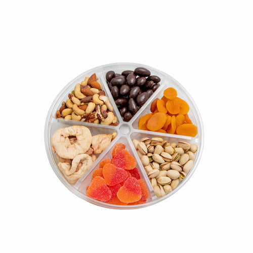A round clear plastic snack box divided into six compartments, filled with assorted nuts, dried fruit, and chocolate-covered pieces, photographed from above on a white background.