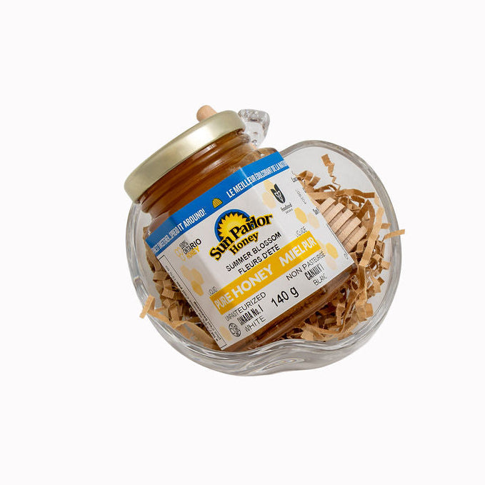 A jar of Sun Parlor Natural Pure Honey Ameba, presented in a clear heart-shaped glass dish filled with shredded paper, on a solid white background.