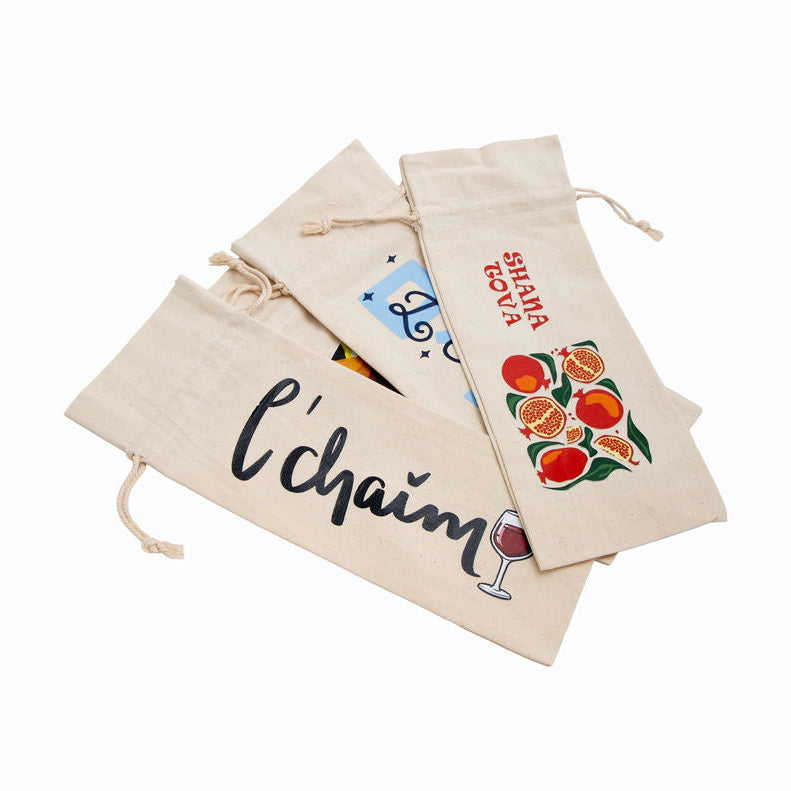 Three beige canvas wine gift bags with drawstring closures, featuring printed designs including black cursive text, red fruit illustrations, and a small blue graphic, arranged on a white background.