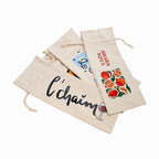 Three beige canvas wine gift bags with drawstring closures, featuring printed designs including black cursive text, red fruit illustrations, and a small blue graphic, arranged on a white background.