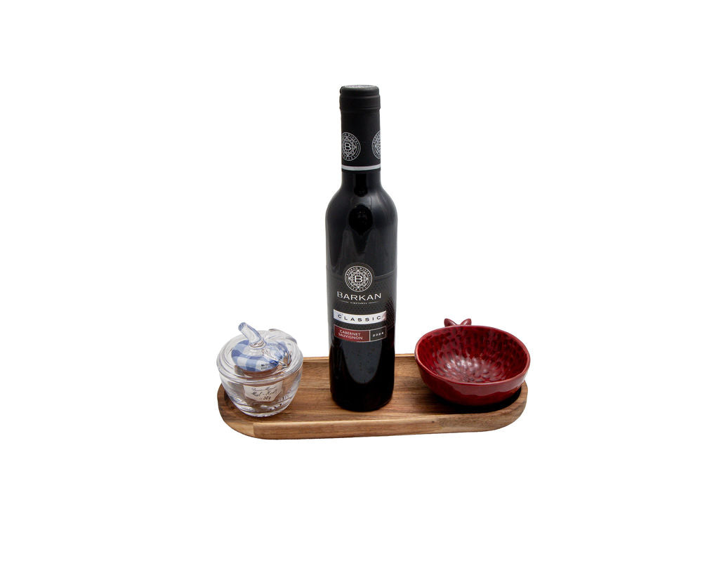 A bottle of Barkan Cabernet Sauvignon centered on a small wooden tray, with a small clear glass container on the left and a textured red bowl on the right, all against a plain white background.