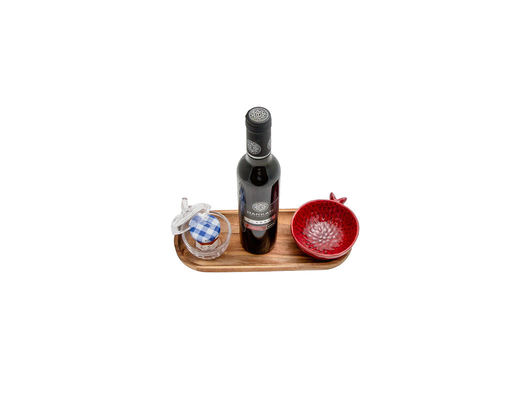 A dark glass bottle with a black cap stands on a small wooden tray, flanked by a small clear glass jar with a blue-and-white checkered lid and a red textured bowl, against a plain white background.