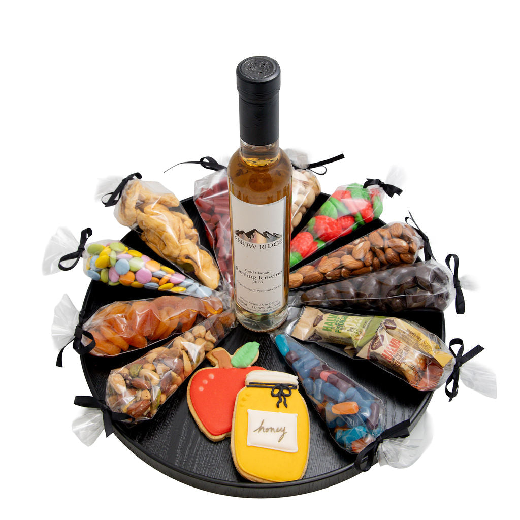 A gourmet gift hamper featuring a bottle of wine centered on a round black tray, surrounded by assorted packaged snacks, candies, nuts, and cookies in clear bags against a white background.
