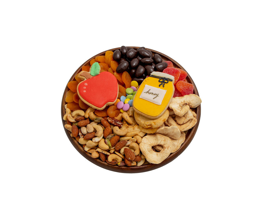 A round wooden board filled with assorted dried fruits, nuts, chocolate-covered pieces, gummy candies, and a red apple-shaped cookie with a green leaf, photographed on a plain white background.