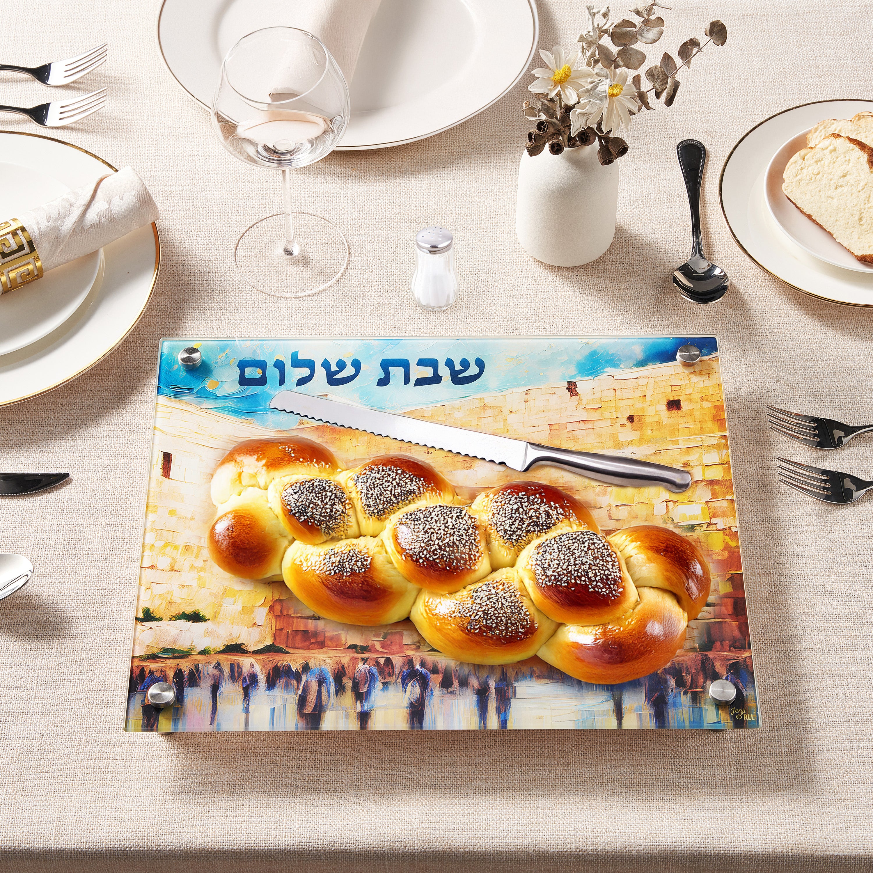 Glass cutting board with Jerusalem design and challah bread on a dining table setting.