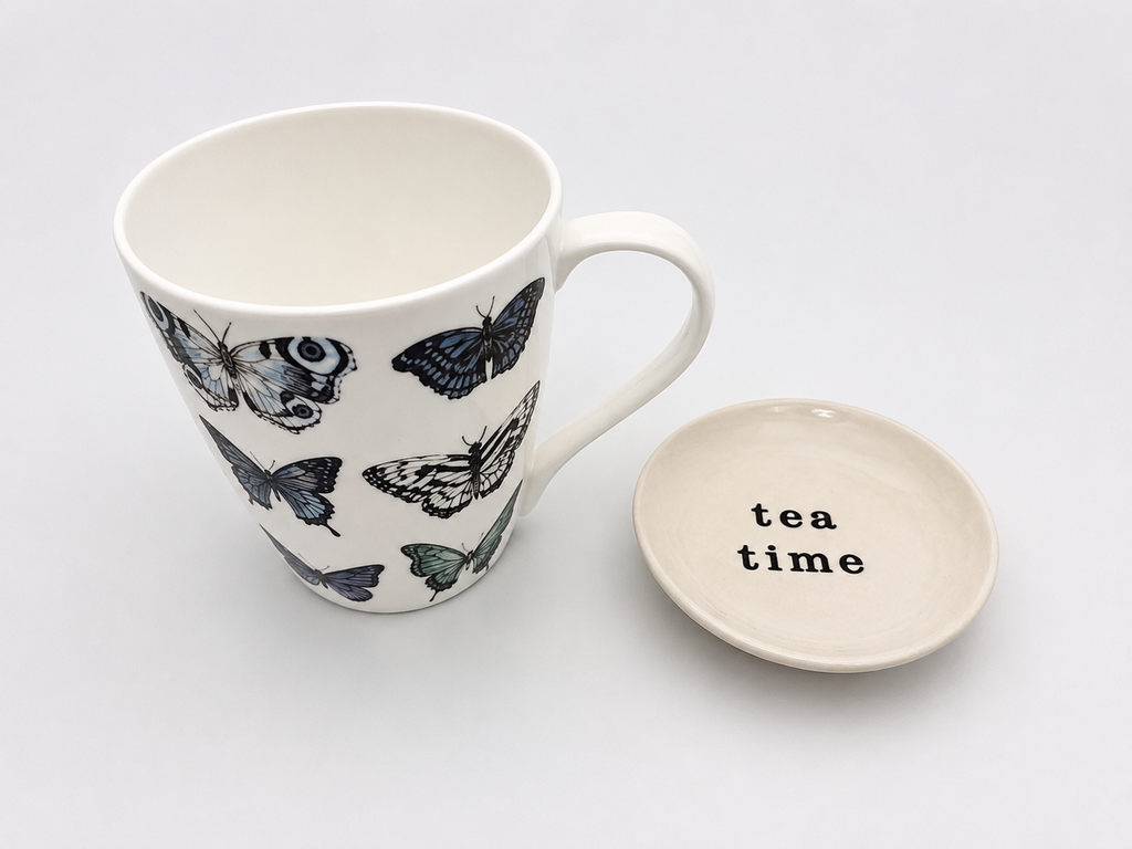 A white ceramic mug decorated with black and blue butterflies, shown next to its matching saucer lid which has the words 'tea time' printed in black.