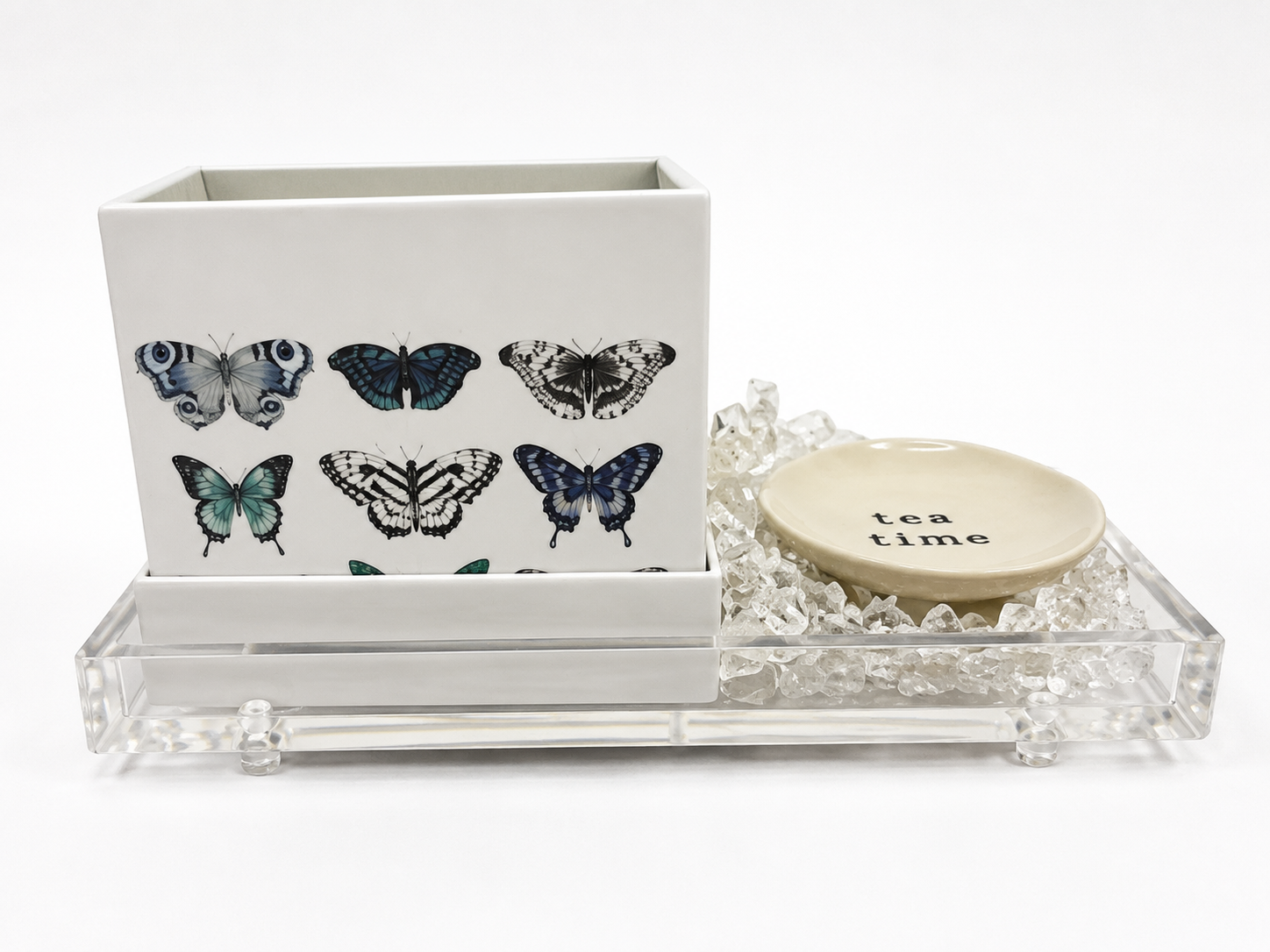 A decorative tea time set featuring a white rectangular box with illustrated butterflies, a small round cream-colored ceramic dish with 'tea time' printed on it, and a clear acrylic tray filled with clear crystal beads.