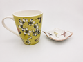 A ceramic mug with a yellow-green body decorated with black and white flowers and small birds, shown next to a matching small saucer on a light gray background.