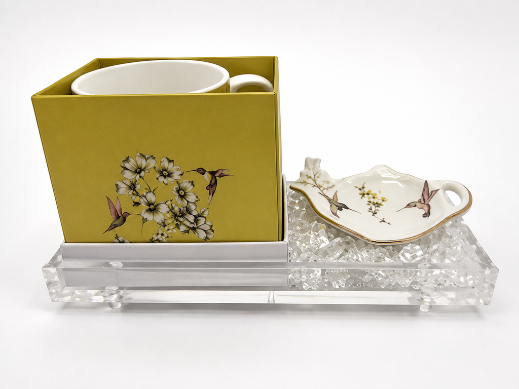 A ceramic mug and matching saucer with floral and hummingbird illustrations, presented in a yellow-green box beside a clear acrylic tray on a white background.